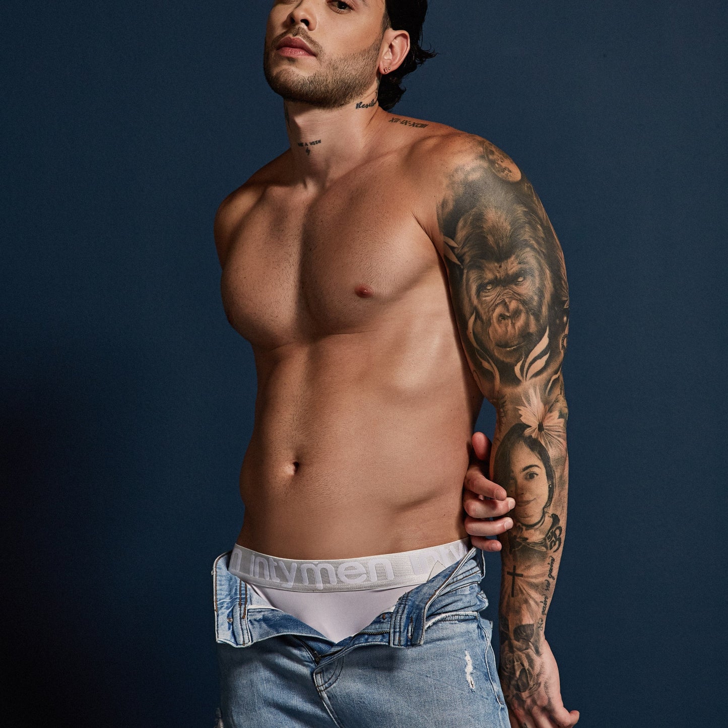 A shirtless, tattooed man confidently poses against a dark backdrop. He wears a black cap, blue jeans, and white Intymen Energy Brief Underwear that enhances his figure. His tattoos, featuring various portraits and designs, highlight his vibrant persona.