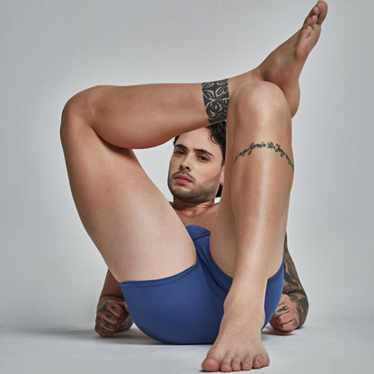 A person in blue Intymen Star Pouch Boxer Trunk poses indoors with bent legs, showcasing arm and thigh tattoos. The soft, stretchy fabric suits their neutral expression and short hair against a plain background.