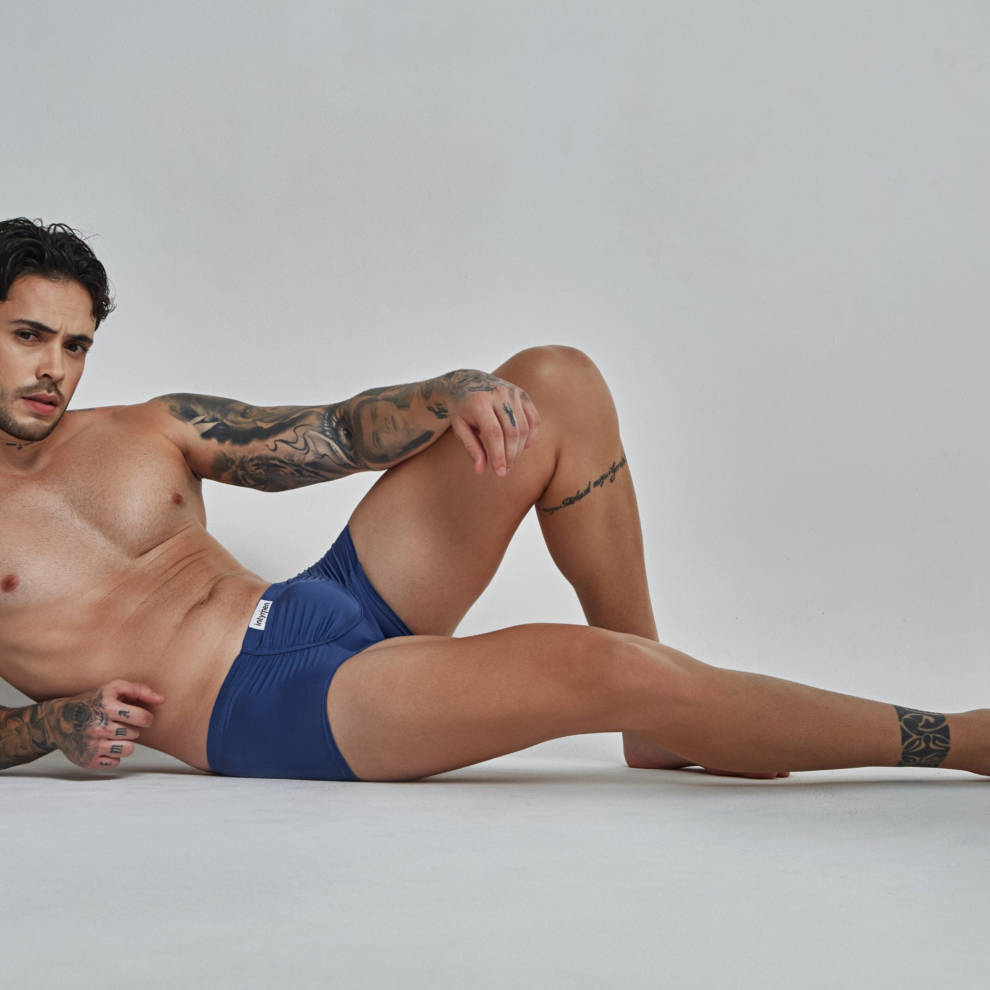 A tattooed man with dark hair poses shirtless, wearing Intymen Star Pouch Boxer Trunk Underwear, made of soft, stretchy fabric. Lying on his side against a plain gray background, he exudes confidence while looking at the camera.
