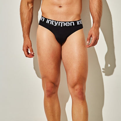 A person stands against a plain background, wearing Intymen Energy Brief Underwear with a white waistband displaying the brand name intymen. Highlighting the luxurious fabric blend and figure-hugging fit, the individual is shown from neck to thighs, emphasizing the sleek design.