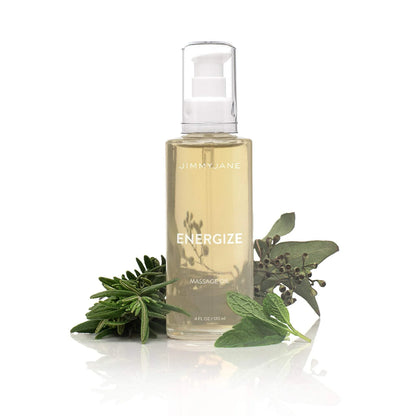A clear pump bottle of Pipedream Products JimmyJane Energize Massage Oil - 4 oz sits beside fresh sage, mint, and rosemary on a white background, emphasizing its revitalizing aromatherapy botanical ingredients.