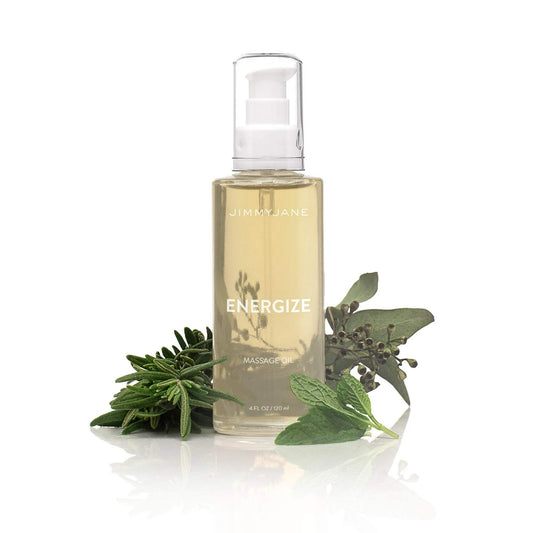 A clear pump bottle of Pipedream Products JimmyJane Energize Massage Oil - 4 oz sits beside fresh sage, mint, and rosemary on a white background, emphasizing its revitalizing aromatherapy botanical ingredients.