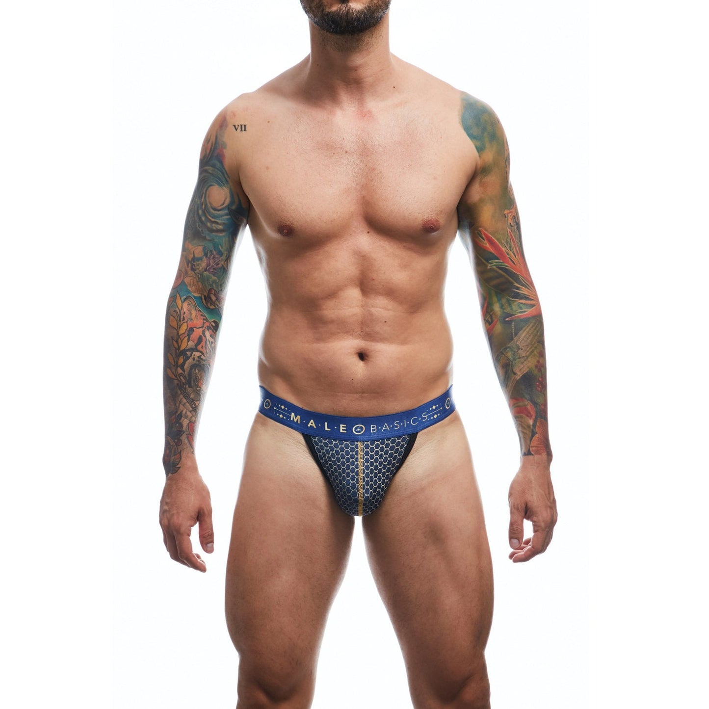A shirtless, tattooed man faces forward in the Malebasics Hipster Jockstrap Andalusia, featuring patterned blue briefs with a low-cut waistband. The luxurious fabric blend delivers comfort and style against the stark white background.