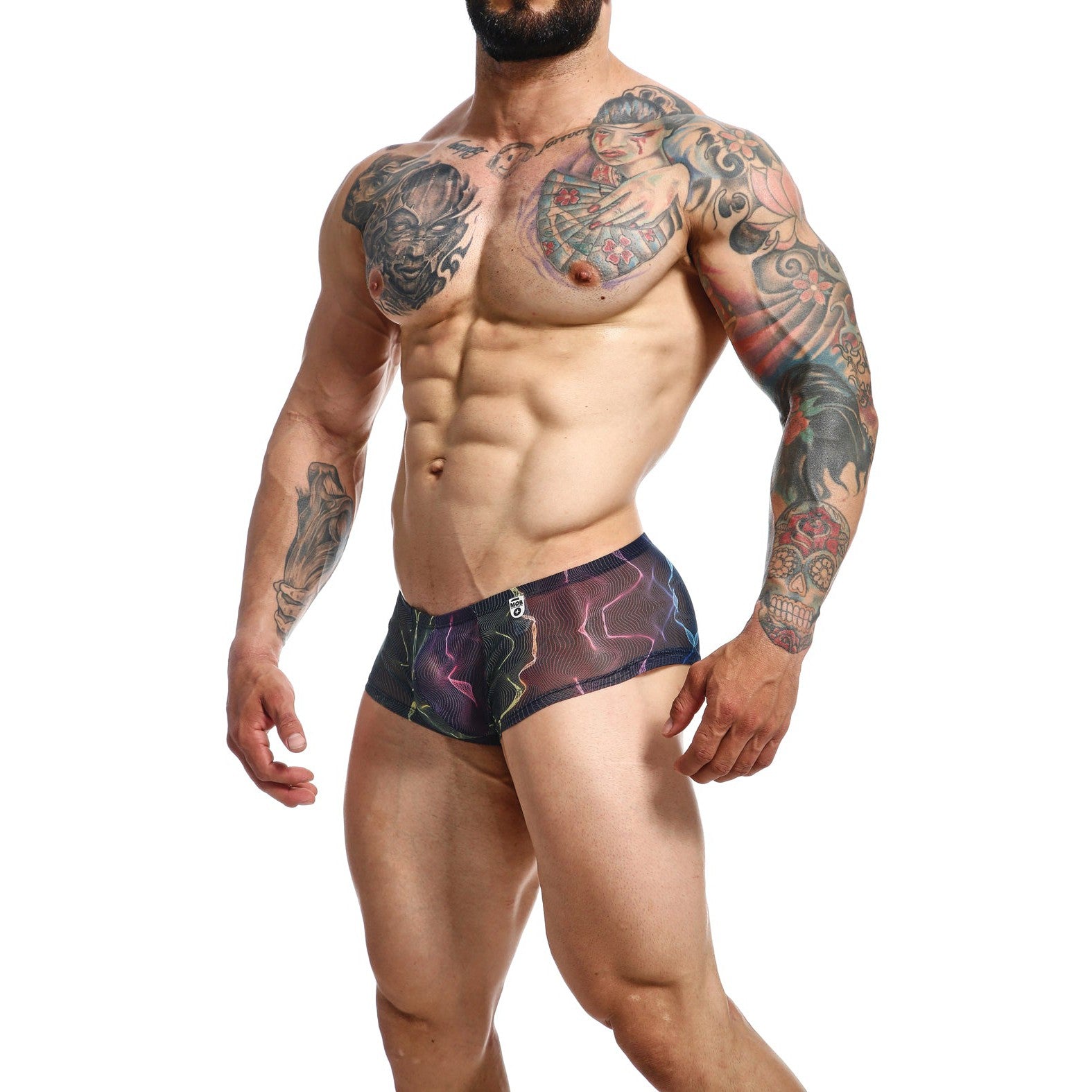 A muscular man models the Malebasics MOB Hip Hugger Boyshort Energy in black, with sheer mesh revealing his arm, chest, and abdomen tattoos of skulls, flowers, and abstract shapes.