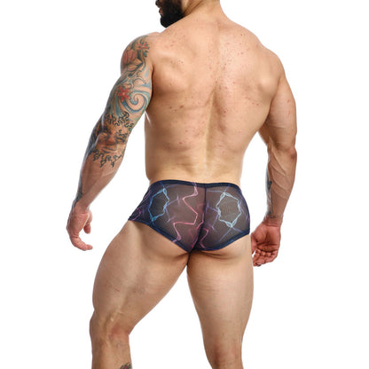 A muscular tattooed man stands slightly turned away wearing Malebasics MOB Mens Hip Hugger Boyshort Energy swim briefs. Small skin blemishes are visible against a plain white background.