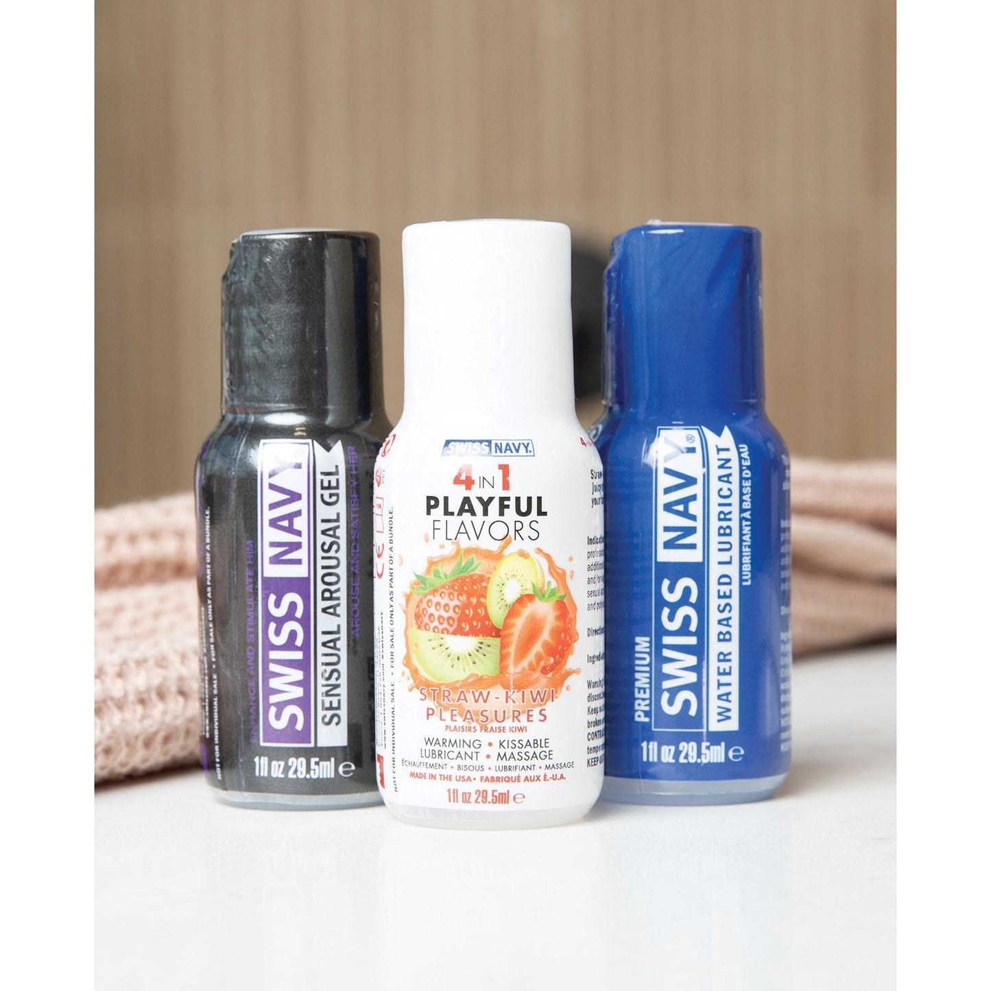 The enticing Swiss Navy Weekend Fun Pack of 3 - 1 oz features Sensual Arousal Gel, a strawberry-kiwi scented 4-in-1 Playful Flavors massage gel, and Premium Water Based Lubricant. These three small bottles rest on a light surface with soft, light brown fabric in the background.