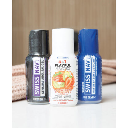 The enticing Swiss Navy Weekend Fun Pack of 3 - 1 oz features Sensual Arousal Gel, a strawberry-kiwi scented 4-in-1 Playful Flavors massage gel, and Premium Water Based Lubricant. These three small bottles rest on a light surface with soft, light brown fabric in the background.