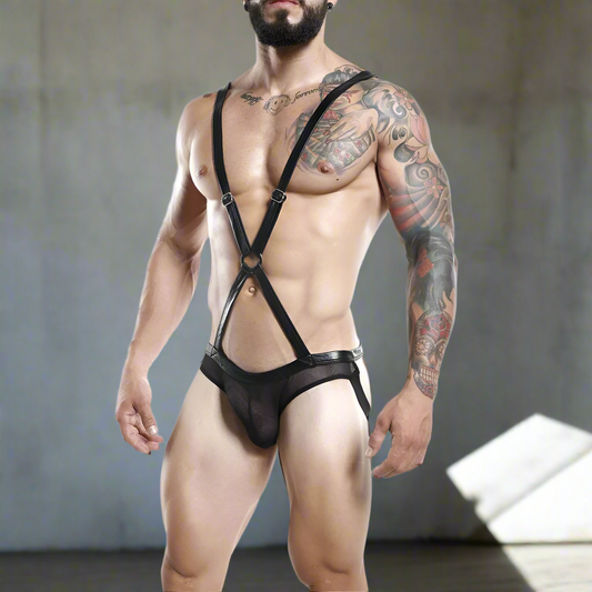 A muscular, tattooed man poses in a strappy Miami Jock Body Suit by Miami Jock, paired with mesh underwear. Set against a neutral industrial background, his face remains partially hidden.