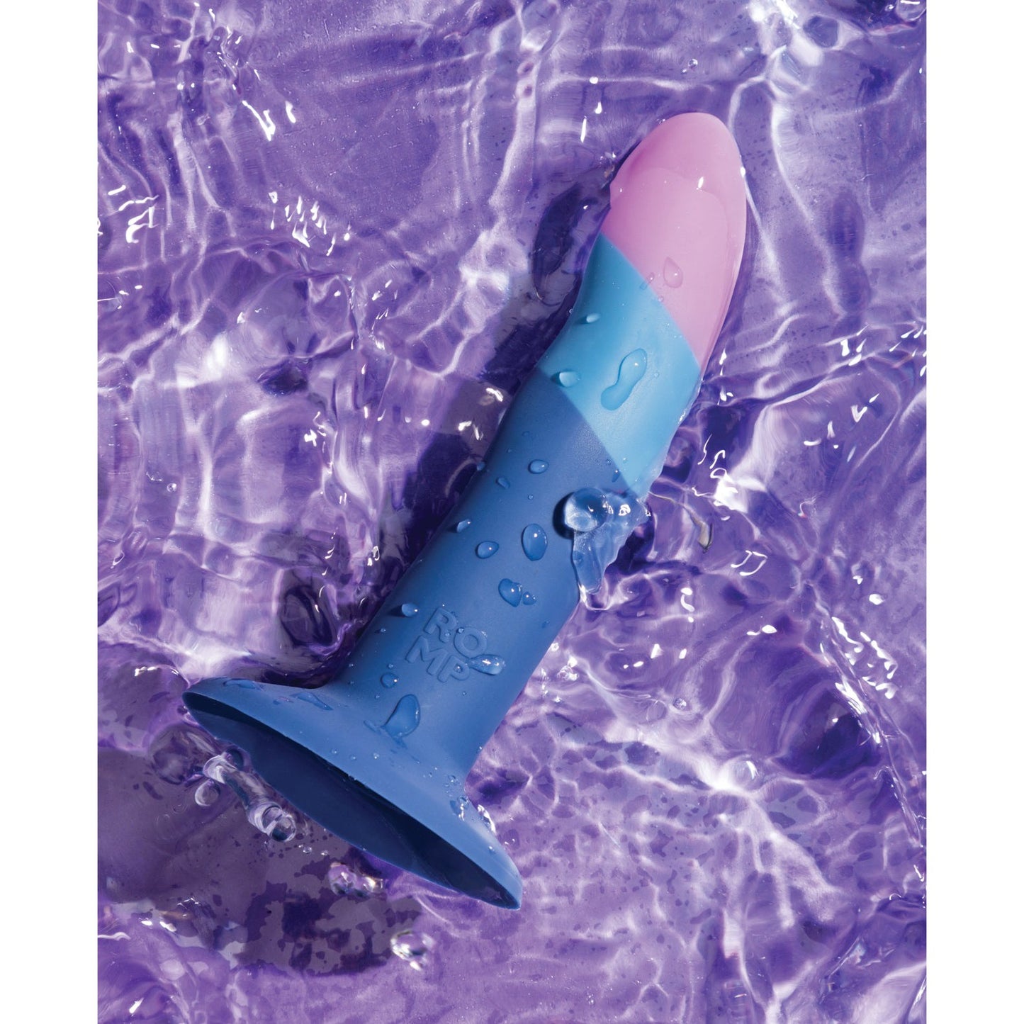 A ROMP Piccolo 3 Color Dildo made from body-safe silicone rests on a wet purple surface. Water droplets accentuate its vibrant blue and pink hues, while the P-spot-friendly design and suction base keep it securely in place.