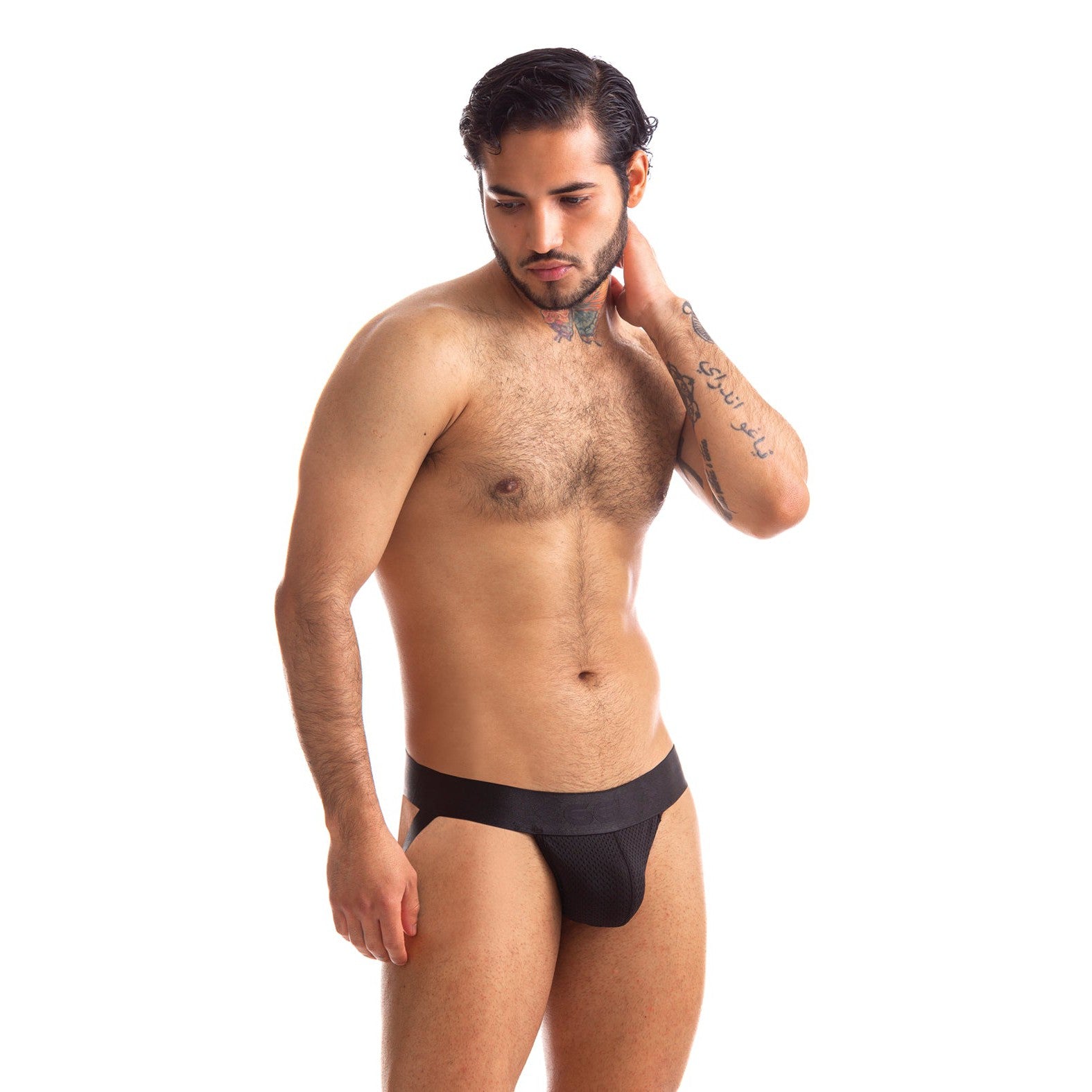 A man with dark hair stands against a white background, wearing the 665 Stealth Jockstrap by 665 inc, featuring a see-through mesh pouch. He has tattoos on his left arm and touches the back of his neck, appearing thoughtful.
