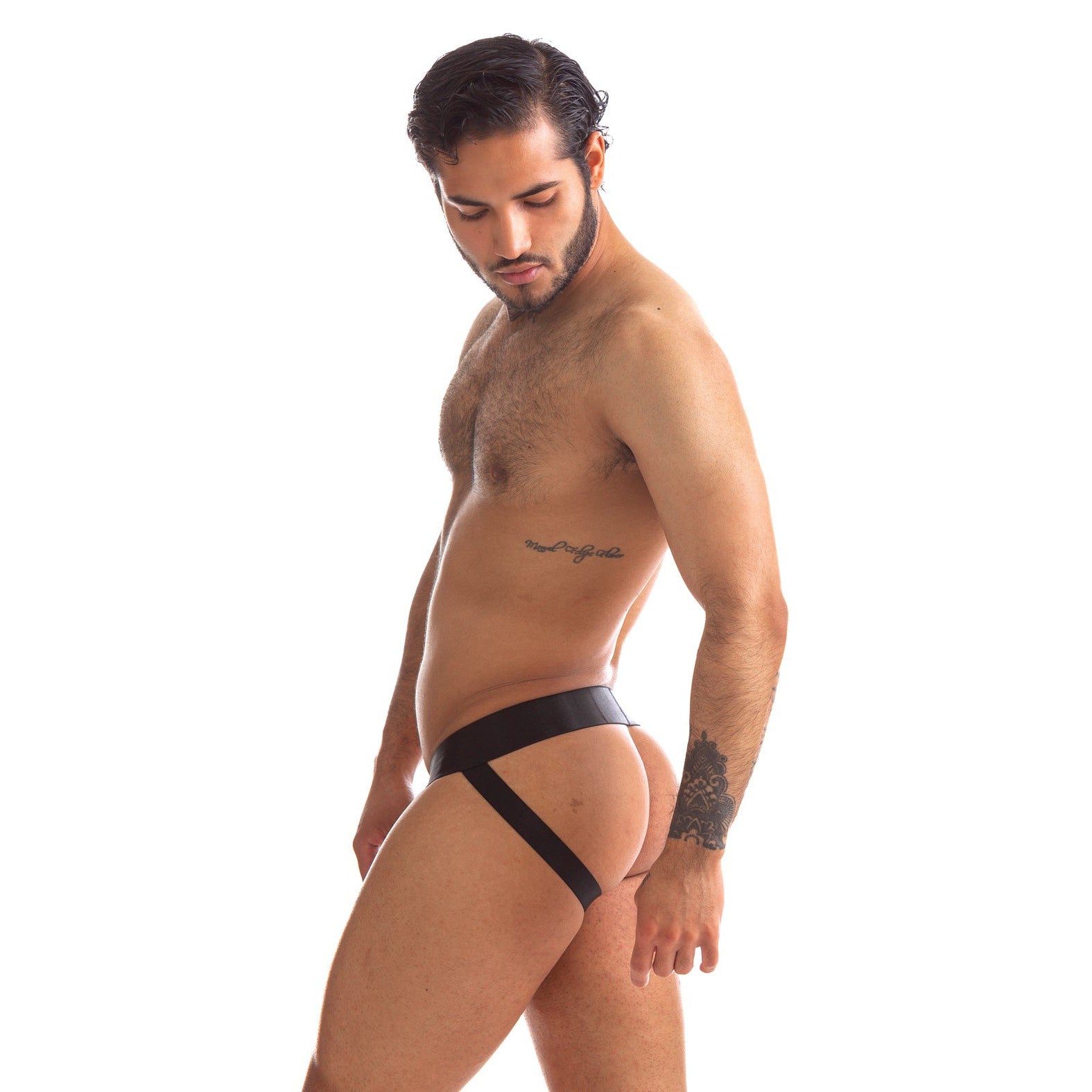 A man with short dark hair confidently poses in the 665 Stealth Jockstrap by 665 inc, featuring a mesh pouch. Set against a white background, he stands sideways with a tattoo on his left forearm, gazing down at the ground.