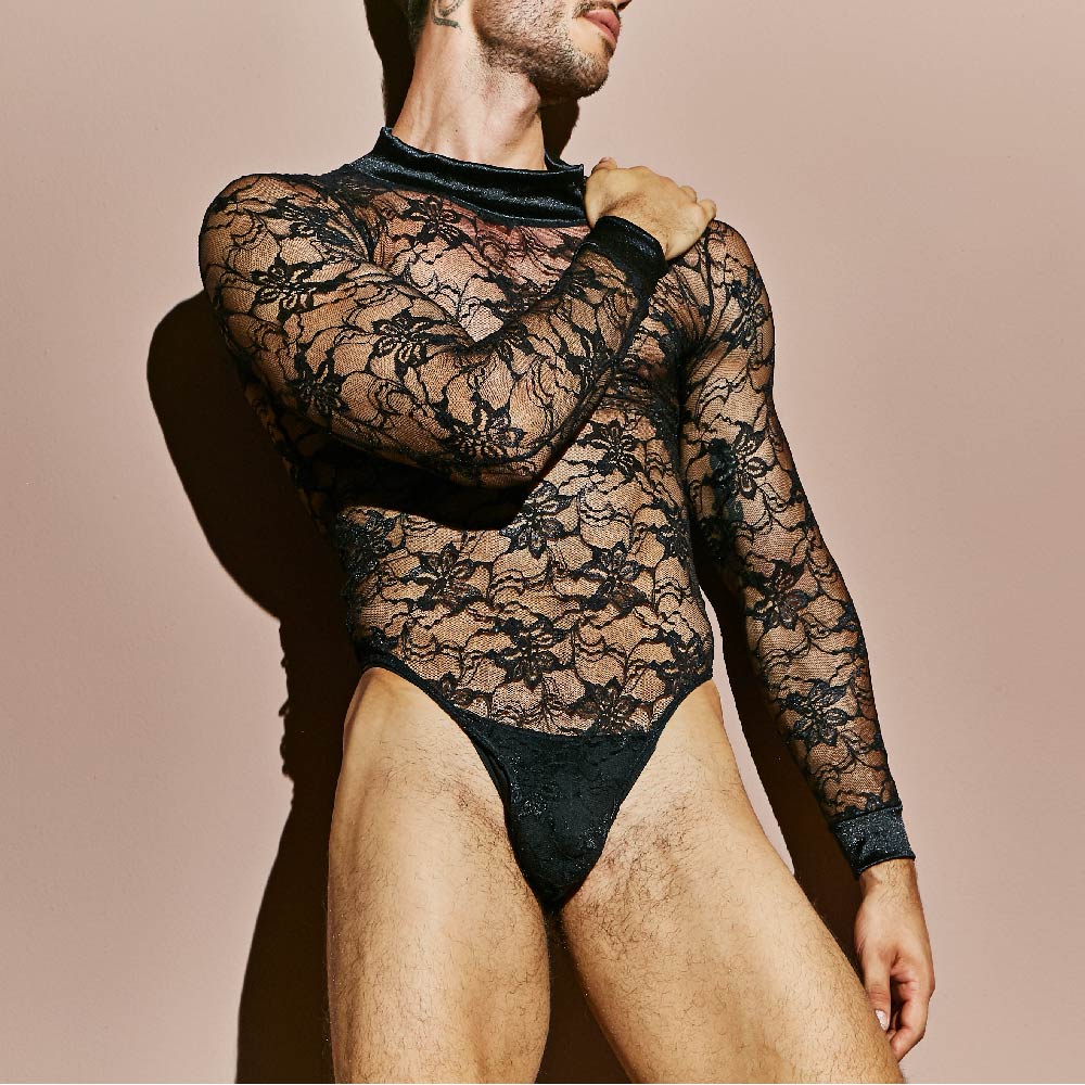 Someone is wearing the Secret Male Mens Lingerie Sexy Bodysuit, featuring alluring black lace with a floral pattern. Against a plain beige background, the long-sleeved suit hugs the body and is slightly sheer. The persons head is turned to the side and not visible.
