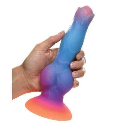 A hand holds the Creature Cocks Space Cock Silicone Alien Dildo, showcasing its glow-in-the-dark, multicolored design. The artistic piece features a wide base and textured surface in shades of blue, purple, pink, and orange.