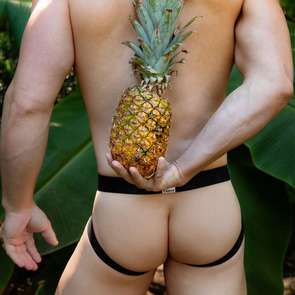 A person wearing an Agacio Thriller Jockstrap stands outdoors with a pineapple behind their back, enhancing the exotic look. Large green leaves frame the scene, adding allure to this premium fabric display.