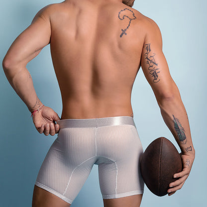 A shirtless man from behind wears white athletic shorts, holding a football in his right hand. His tattooed arms add an edge, and beneath the shorts, Agacio Scuba Boxer Trunks enhance the look against a plain blue wall.
