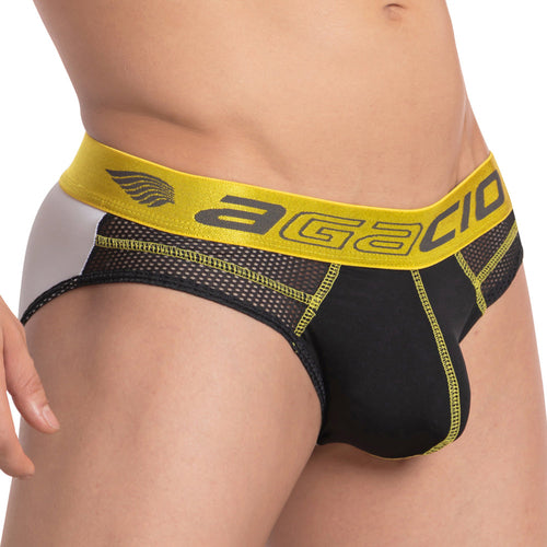 The Agacio The Charged Brief is showcased in a close-up of a persons midsection, highlighting a classy shiny waistband in vibrant yellow. The brand name Agacio stands out prominently, while breathable mesh detailing on the sides enhances the sophistication of the black and white design.
