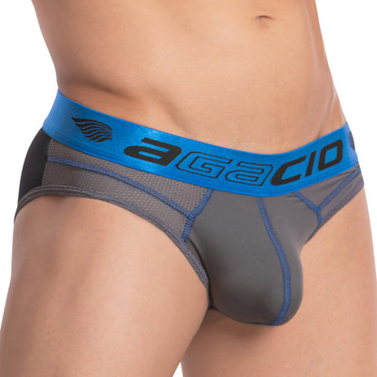 The image displays a person in Agacio The Charged Brief, featuring a gray and black design with a shiny blue waistband boldly displaying Agacio. The fabric is smooth, fitted, and includes breathable mesh for enhanced comfort.
