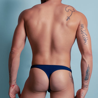 A man, tattooed and facing away from the camera, wears an Agacio Deep Sea Thong in dark blue against a light blue backdrop. The thongs airy fabric offers both comfort and style.