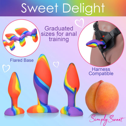 The Curve Toys Simply Sweet Rainbow Silicone Butt Plug Set by Curve Novelties features three vibrant, body-safe silicone plugs in graduated sizes with flared bases. Great for anal training and harness-compatible, the image includes a peach emoji with Sweet Delight and SimplySweet text.