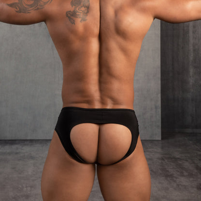 In a concrete-walled room, someone with a muscular back showcases the bold design of Daddys Seductive Gay Jockstrap in black, revealing tattoos on their shoulders.