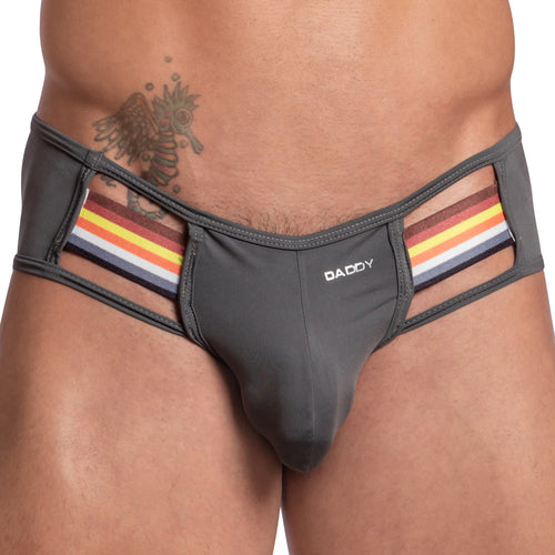 A person showcases a seductive Daddy jockstrap with a bold design—unique gray fabric with rainbow stripes and cut-out details. The Daddy brand is proudly displayed, complementing a winged creature tattoo on their torso.