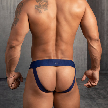 A muscular individual is seen from behind, showcasing the Daddy Wide Waistband Jockstrap in blue, with a tattoo on their left arm against a plain gray wall.