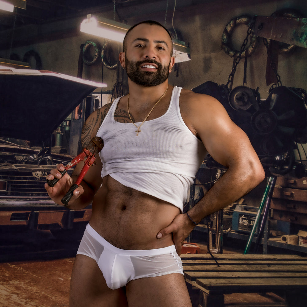 A muscular, bearded man in a white tank top and sheer boxer trunks stands smiling in a workshop filled with tools, car parts, and machinery. He exudes rugged charm while wearing Daddy Enhanced Pouch Boxer Trunk for breathable comfort.