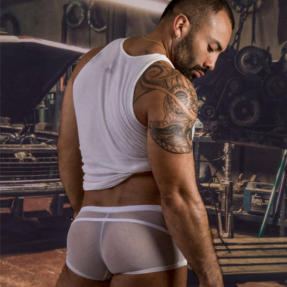 A man with a tattooed arm wears a white tank top and see-through Daddy Enhanced Pouch Boxer Trunks made by Daddy, standing in a workshop filled with tools and machinery.
