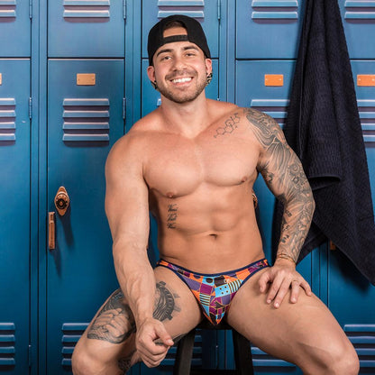 A muscular man, smiling on a bench in a locker room, wears the colorful Daniel Alexander Limited Ass Kini Thong and a black cap. The blue lockers highlight the sleek and stretchy waistband of his unique outfit.