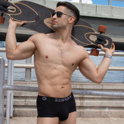A shirtless man in black Edipous Bi-Color Sports Boxer Trunk and sunglasses stands outdoors with a longboard behind his neck, a bridge and water as the backdrop. Hes wearing a necklace, and his boxer trunk has a supportive pouch for added comfort.