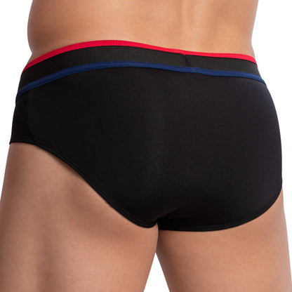 A person wearing the Edipous Munich Brief, a stylish mens bikini featuring black briefs with red and blue waistband stripes. The low-rise design provides a sleek fit, shown from the waist down in the back view.