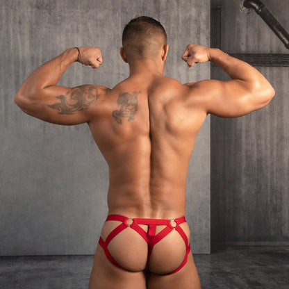 A muscular person stands with their back to the camera, flexing in Good Devils bold red Jockstraps featuring criss-cross straps and a supportive pouch, revealing their back and legs. The background is a gray concrete wall with exposed pipes.