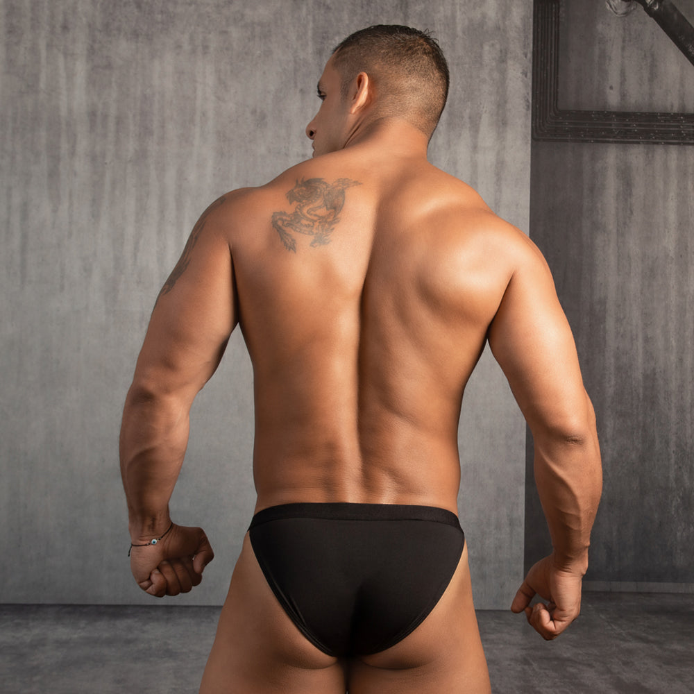 A person with short hair faces away, displaying muscular back and arms. They wear Good Devil Bikinis Steel Ring Waistband in black, with a scorpion tattoo on their right shoulder. The minimalist design complements the concrete room with a metal beam in the corner.