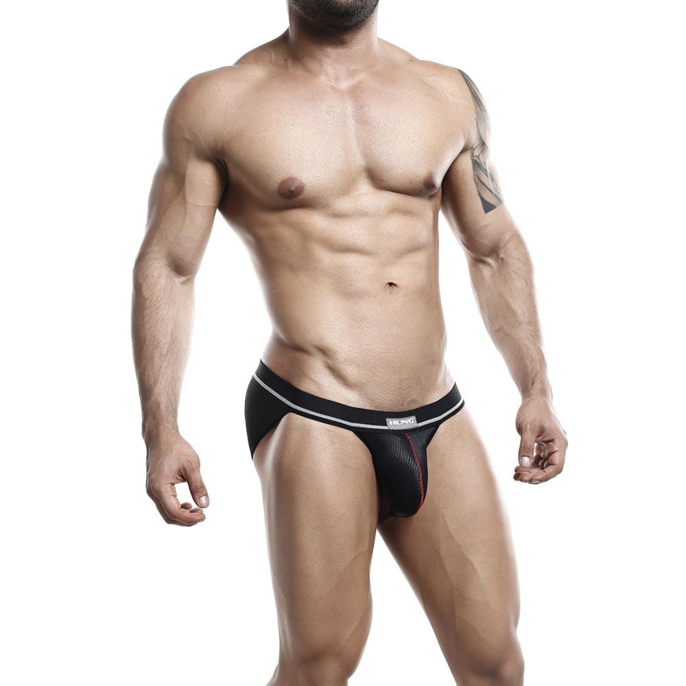 A muscular man models the Hung Jockstrap from Hung, a breathable black and red design highlighting his athletic physique. He poses against a white background, arms relaxed by his sides, displaying a tattoo on his right arm.