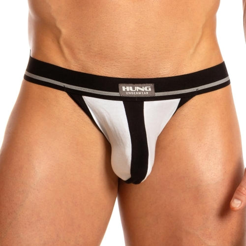 A person models a bold design jockstrap with supportive elastic straps against a plain background. The waistband prominently displays the brand Hung. The Hung Dong Jock features a central black stripe flanked by white fabric.