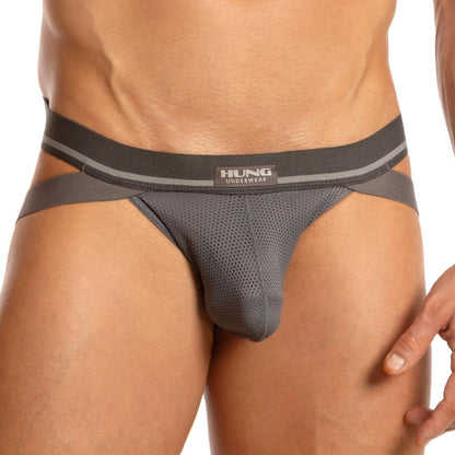 A close-up shows someone in gray mesh Hung Big Boy Jock underwear with a black waistband and side straps. The Hung brand is visible, showcasing its spacious pouch design. The premium fabric blend highlights the persons lower body and hand.