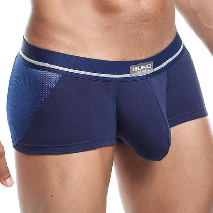 A person wears dark blue Hung Clash Trunk boxer briefs by Hung, featuring a gray waistband with text and mesh side panels. The contour pouch design ensures comfort. Their hand is partly visible on the left against a plain white background.
