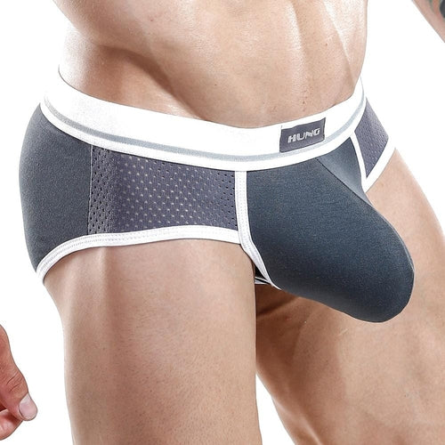 A close-up shows someone in Hung Briefs by Hung, featuring dark gray and white colors. Breathable mesh side panels increase airflow, and a functional pouch provides extra support. The white waistband displays the logo in the center, exposing the wearers torso and upper legs.
