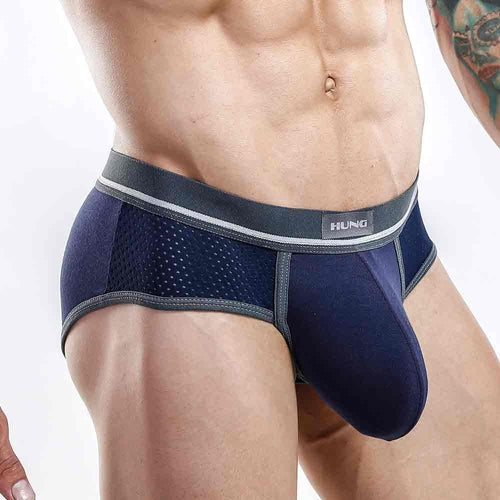 A person in navy blue and gray Hung Briefs by Hung stands against a plain backdrop. The briefs, featuring a branded waistband and mesh sides for breathability, ensure comfort. A tattoo is partially visible on their arm.