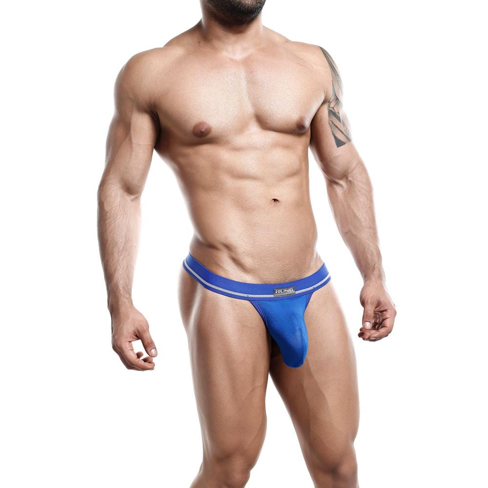 A muscular model with a tattoo on their upper left arm confidently poses in blue Hung Accessories underwear by Hung. Theyre slightly turned to the side, arms relaxed by their sides, showcasing the enhancing pouch design.