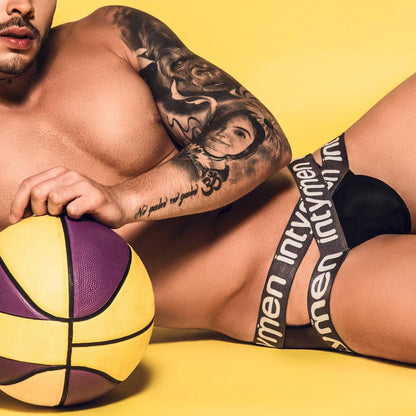 A tattooed man lies on a yellow background holding a vibrant basketball, wearing Intymen Cross Jockstrap with its bold design and prominent brand name displayed on the waistband.