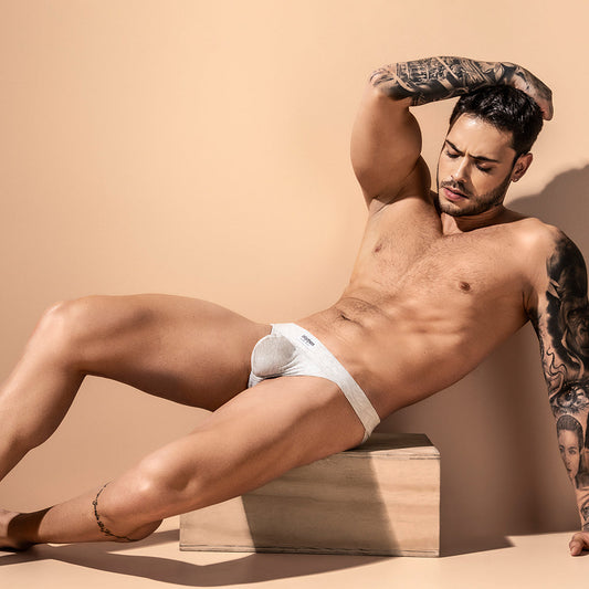 A tattooed, muscular man reclines on a wooden box against a beige backdrop, showcasing the Intymen Loving Jockstraps supportive thick waistband. His eyes are closed, one arm raised over his head, highlighting his toned form in a cotton-spandex blend by Intymen.