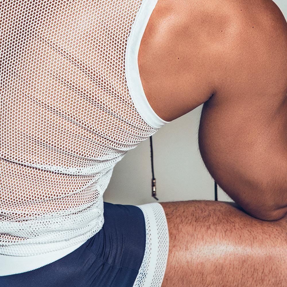 A person is sitting sideways wearing the Intymen Quads Trunk by Intymen, featuring a white mesh tank top and black shorts that fit impeccably. The image zooms in on the upper body and arm, emphasizing the premium fabric texture against the skin.