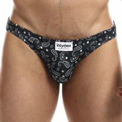 A close-up shows a person wearing Intymen Sexy Boy Paisley Bikini briefs featuring an intricate black paisley pattern. The waistband displays the brand name Intymen, and the Royal Paisley Print stands out beautifully against the white background.