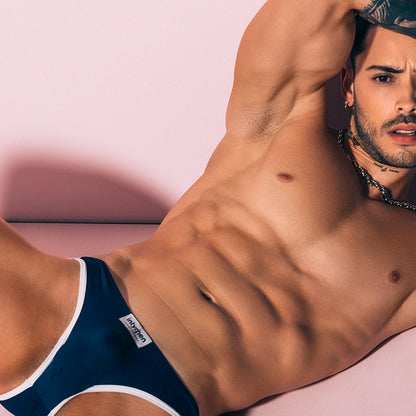 A shirtless man showcases his muscles on a pink couch wearing navy blue Intymen Veneto Bikini underwear with white trim and label. The polyamide-spandex fabric is breathable, sheer, highlighting his tattoos as he raises an arm and wears a chain necklace.