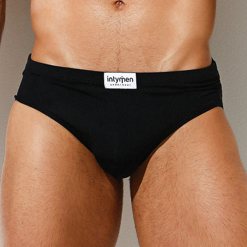 A close-up image shows someone wearing Intymen Amore Brief in black, with the label visible on the waistband. The briefs provide contoured pouch support, and their luxurious fabric contrasts elegantly against a neutral wall background.