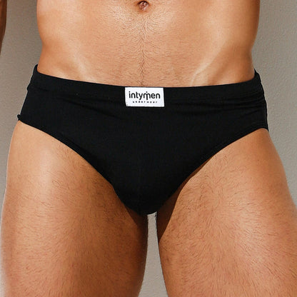 A close-up image shows someone wearing Intymen Amore Brief in black, with the label visible on the waistband. The briefs provide contoured pouch support, and their luxurious fabric contrasts elegantly against a neutral wall background.