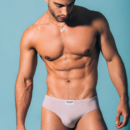 A muscular man models Intymens Amore Brief in light gray against a blue background, wearing a gold pendant necklace. His toned abs and partially visible left arm highlight the luxurious fabrics contoured pouch support, enhancing his confident stance.
