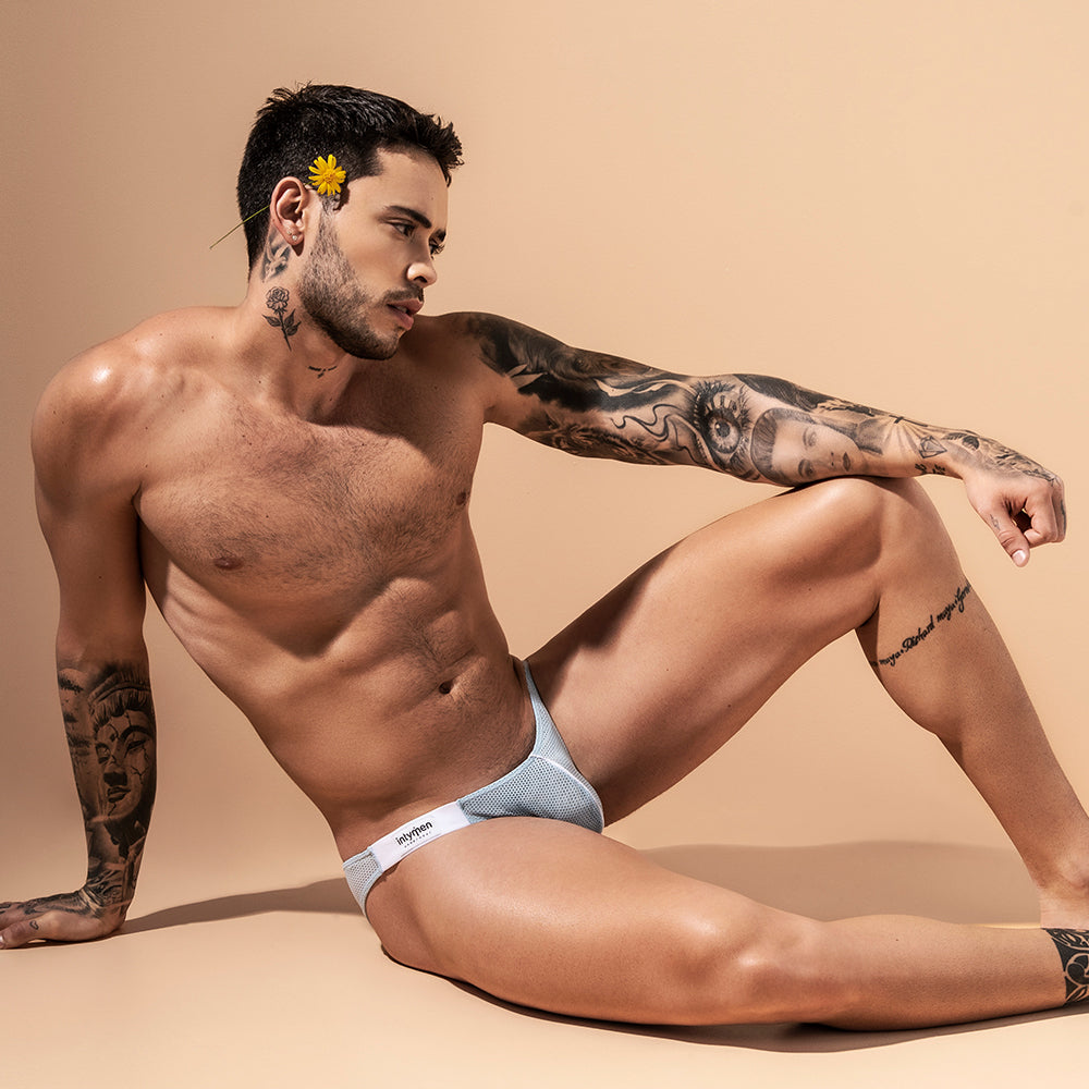 A tattooed man lounges on a beige surface in light gray Intymen Fancy Briefs, which are low-waisted. A yellow flower is tucked behind his ear as he gazes to the side.