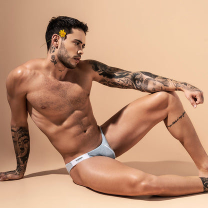 A tattooed man lounges on a beige surface in light gray Intymen Fancy Briefs, which are low-waisted. A yellow flower is tucked behind his ear as he gazes to the side.